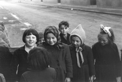 Black and white photograph of a group of smiling children in hats and coats on a street