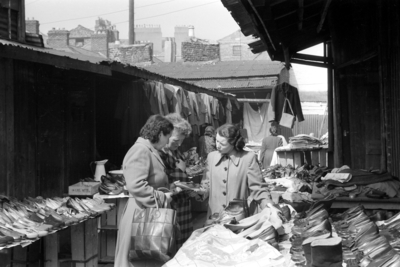 Black and white photograph of women browsing shoes and clothes at a covered market stall