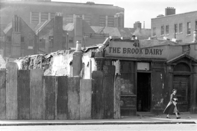 Black and white photograph of a woman walking past a hoarding and the derelict front of The Brook Dairy