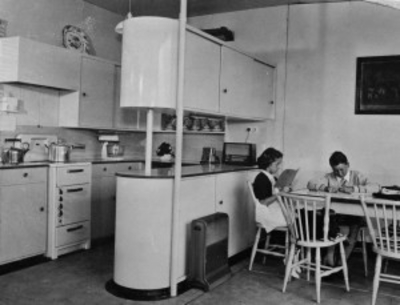 Black and white photograph of a fitted kitchen with a woman and a child at the table