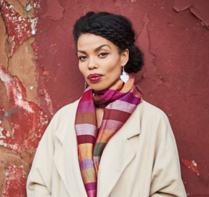 Emma Dabiri, Explore Your Archive 2020 ambassador, in a cream coat and striped scarf against a red wall