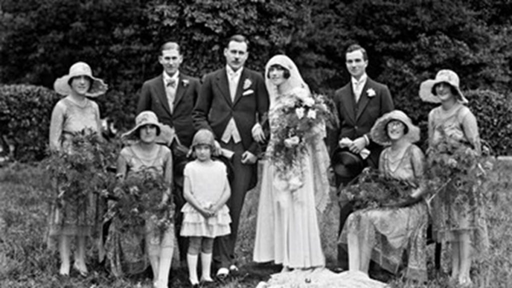 Black and white photograph of a wedding party, with the bride and groom, bridesmaids in wide hats and a young flower girl