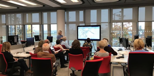 A workshop group seated at computers in a reading room, watching a presenter beside a screen