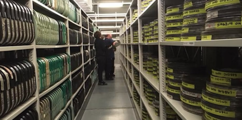 Two people standing in a long aisle of shelving stacked with film reels