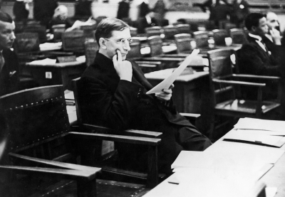 Black and white photograph of a delegate seated at a desk in an assembly hall, reading a document