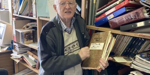 A man holding up a bound volume in a study lined with shelves of books and papers